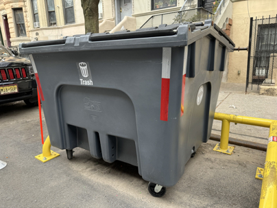 Close-up of a large lidded rolling curbside trash container with the word "Trash" and a garbage can symbol on a residential street in New York City